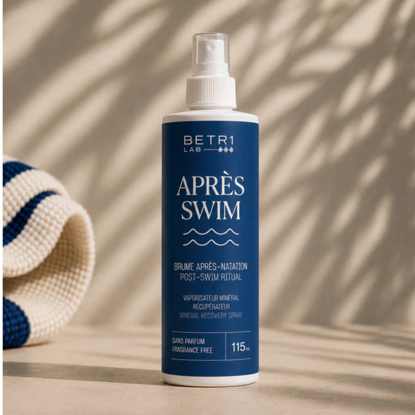 A bottle of BETR1 LAB Après Swim fragrance-free chlorine detox mineral recovery spray, featuring a deep blue label and white spray nozzle, set against a warm beige background with leaf shadows and a rolled towel on the side.